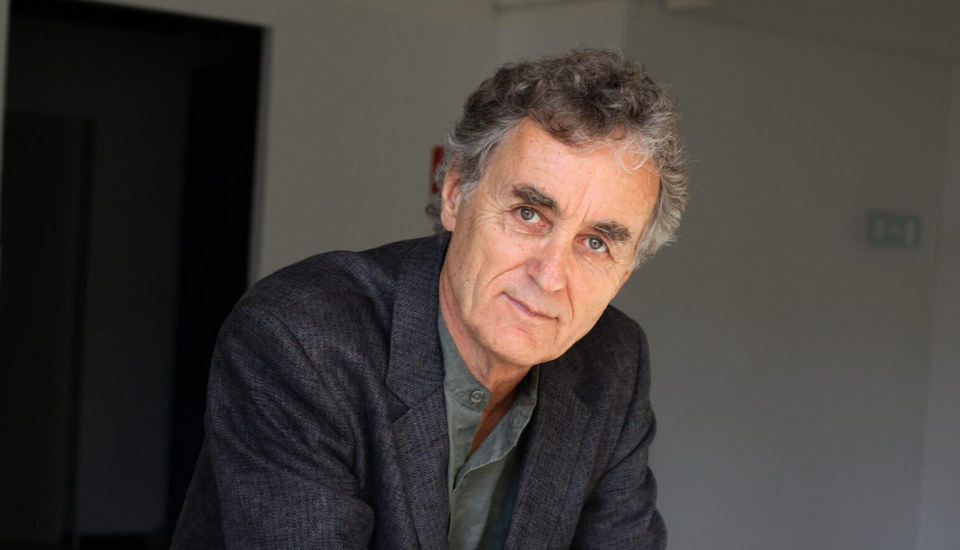A man with gray hair and a thoughtful expression is wearing a dark blazer over a shirt, leaning slightly forward, in a softly lit indoor setting with a neutral background.