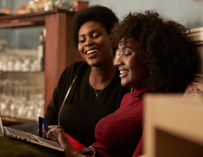 Two people sitting together on a couch, smiling at a laptop screen. One person is holding a credit card. The background has a wooden cabinet with glassware. The scene suggests they might be shopping online or browsing together.