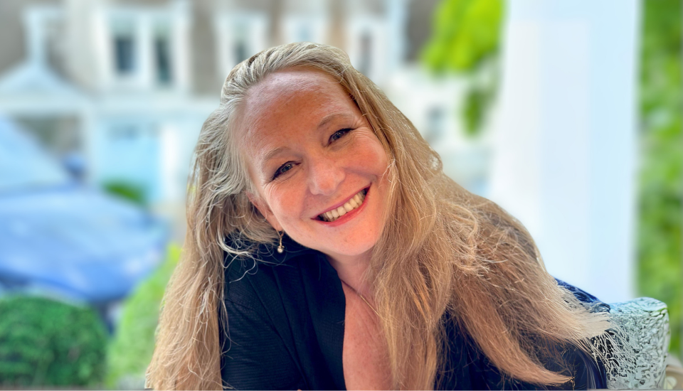 A woman with long blonde hair smiles warmly at the camera. She is wearing a black top. The background is blurred, showing greenery and the outline of a house.