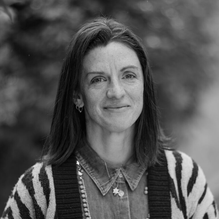 A woman with shoulder-length dark hair, wearing a patterned sweater over a button-up shirt and necklaces, smiles softly while standing outdoors at a sustainability event, with a blurred leafy background. Black and white photo.