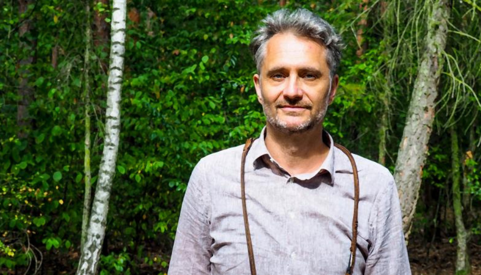 A man with gray hair and a beard stands outdoors in front of lush green trees. He is wearing a light gray button-up shirt with brown suspenders. The setting appears to be a forested area.