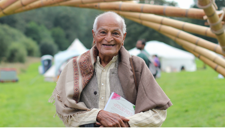 Elderly man with gray hair smiles warmly while holding a booklet in a green outdoor setting. He wears a beige shirt and a patterned shawl. Wooden structure and tents are visible in the background.