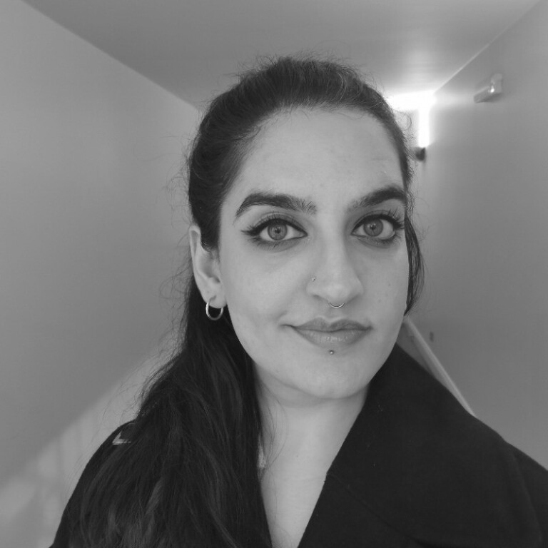 A woman with long dark hair, hoop earrings, and facial piercings stands in a well-lit hallway at the RSA and LGBTQ+ Community Centre in conversation with Sharan Dhaliwal, looking directly at the camera with a neutral expression. The photo is in black and white.
