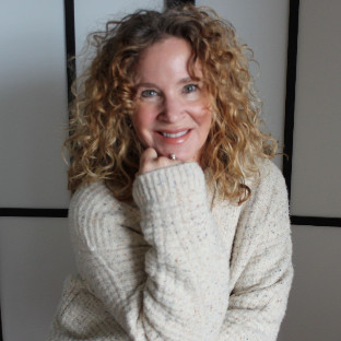 A woman with curly blonde hair and a cozy cream-colored sweater smiles warmly at the camera, resting her chin on her hand. She stands in front of a light background with black lines, radiating a sense of community and connection.