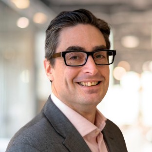 A man wearing dark glasses, a gray suit, and a light pink shirt smiles at the camera, ready to connect. The background is softly blurred, suggesting an indoor office setting with bright lights and a vibrant sense of community.