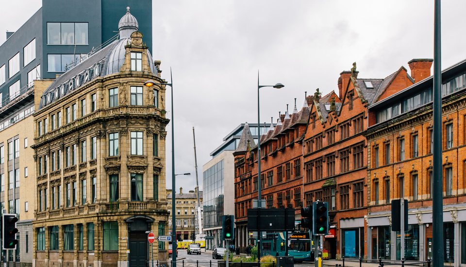 A city street scene with historic stone and red-brick buildings alongside modern glass structures, under a cloudy sky. Traffic lights, street lamps, and a few vehicles are visible on the road.
