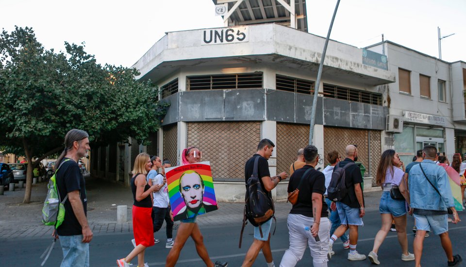 A group of people walk past a worn building with "UN65" painted on it. One person carries a rainbow flag featuring a face. Trees and other buildings are in the background.
