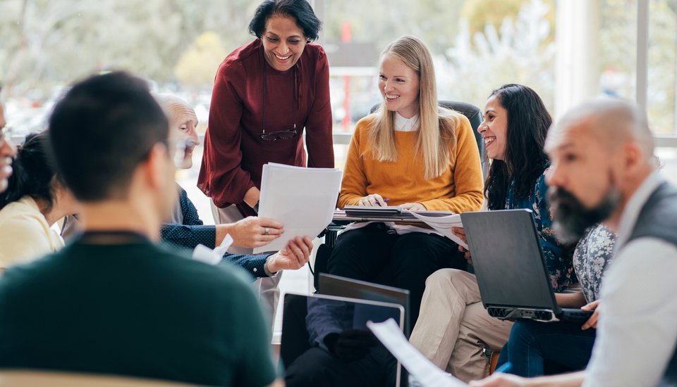 A diverse group of colleagues sit in a circle, discussing papers and using laptops. They are smiling and engaged in conversation, suggesting a collaborative and positive meeting in a bright, modern office space.