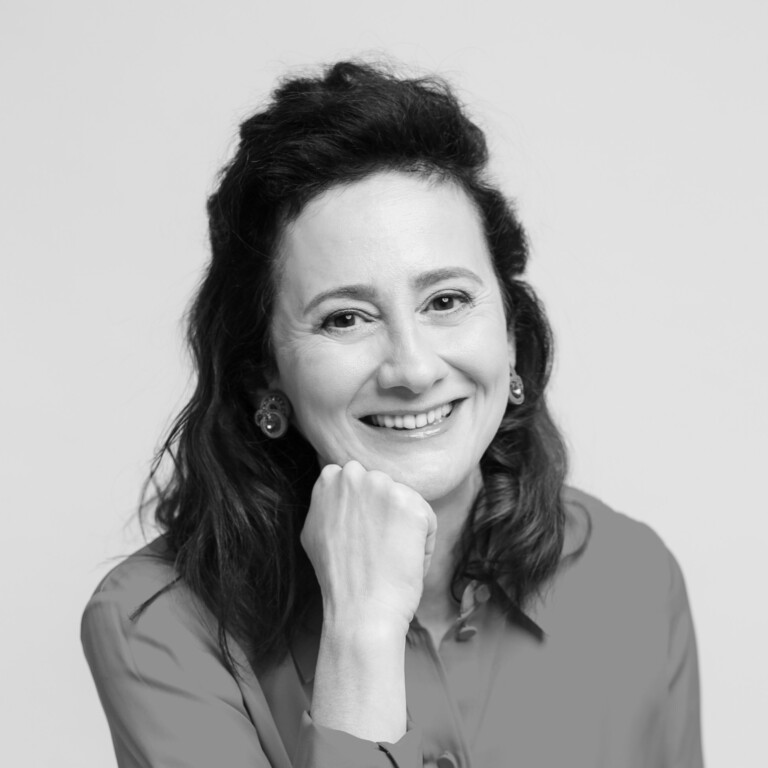 A woman with dark, wavy hair smiles at the camera, resting her chin on her hand. She is wearing a button-up shirt and earrings. The photo is in black and white with a plain background.