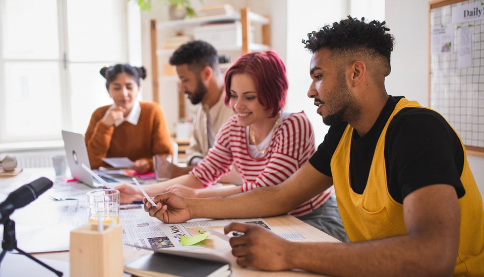 Four young adults sit around a wooden table, collaborating on work with laptops, papers, and pens. They appear focused and engaged, with a bright, modern office space in the background.