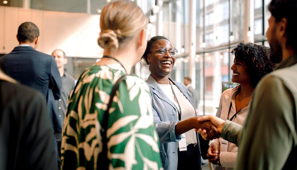 A group of professionals stands in a bright, modern office lobby, smiling and shaking hands while engaging in friendly conversation at a networking event.