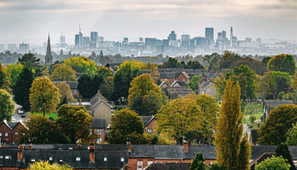 A view of a suburban neighborhood with many trees in early autumn, houses with dark roofs, and a distant city skyline under a cloudy sky—a scene reflecting the balanced communities championed by the RSA Inclusive Growth Commission.