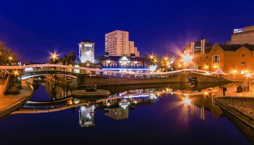 Night view of a canal in a city featuring bridges and illuminated buildings symbolizing inclusive growth, with their bright reflections shimmering on the calm water beneath a deep blue sky.