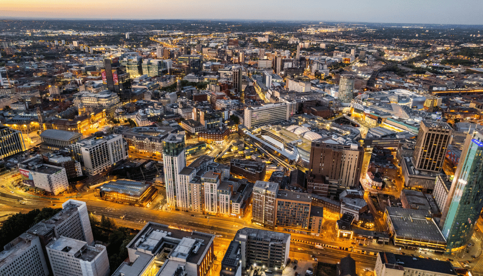 Aerial view of a city at dusk, with buildings, streets, and lights illuminating the urban landscape as daylight fades; a scene reflecting the vision of inclusive growth championed by the East Midlands Inclusive Growth Commission.