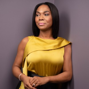 A woman with straight black hair wearing a sleeveless yellow satin top stands against a gray wall, looking thoughtfully to the side as if reflecting on community and ways to connect with others.
