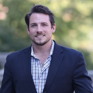 A man with short brown hair and a beard, wearing a dark blazer over a plaid shirt, stands outdoors smiling, ready to connect with his community against a blurred green background.