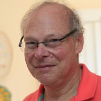 A middle-aged man with glasses and thinning hair smiles slightly, wearing a coral-colored shirt. The softly lit background, featuring a white door and artwork, hints at his warmth and desire to connect with his community.