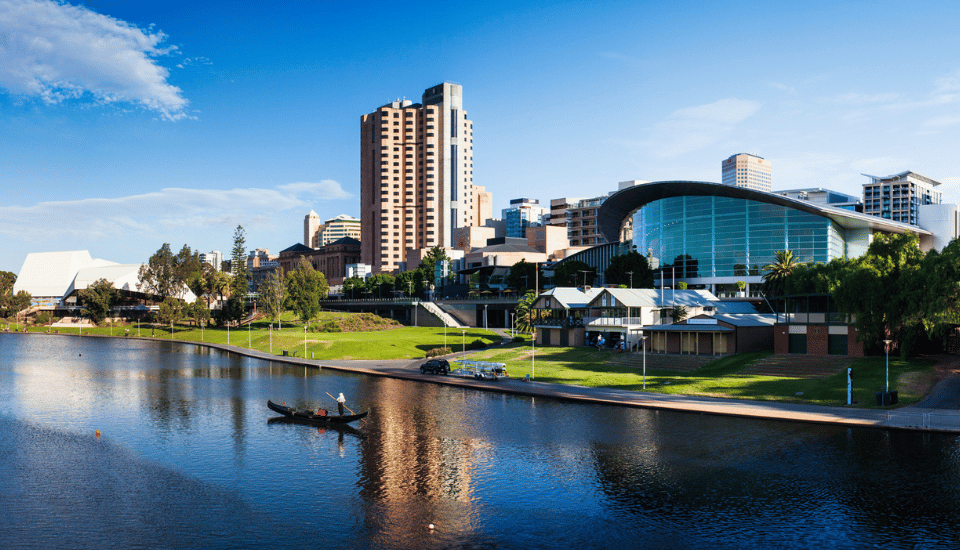 A city skyline with modern and high-rise buildings beside a river, a small boat on the water, and clear blue sky in the background. Green trees and a grassy area line the riverbank.