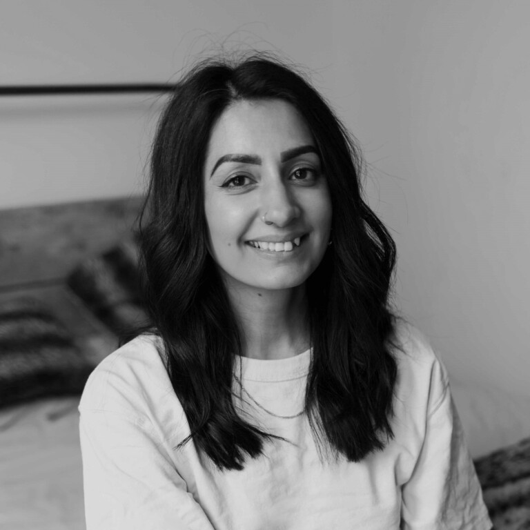 Black and white photo of a young woman with wavy dark hair, smiling softly. She is wearing a light-colored top and is sitting indoors, with a bed and pillows visible in the background.