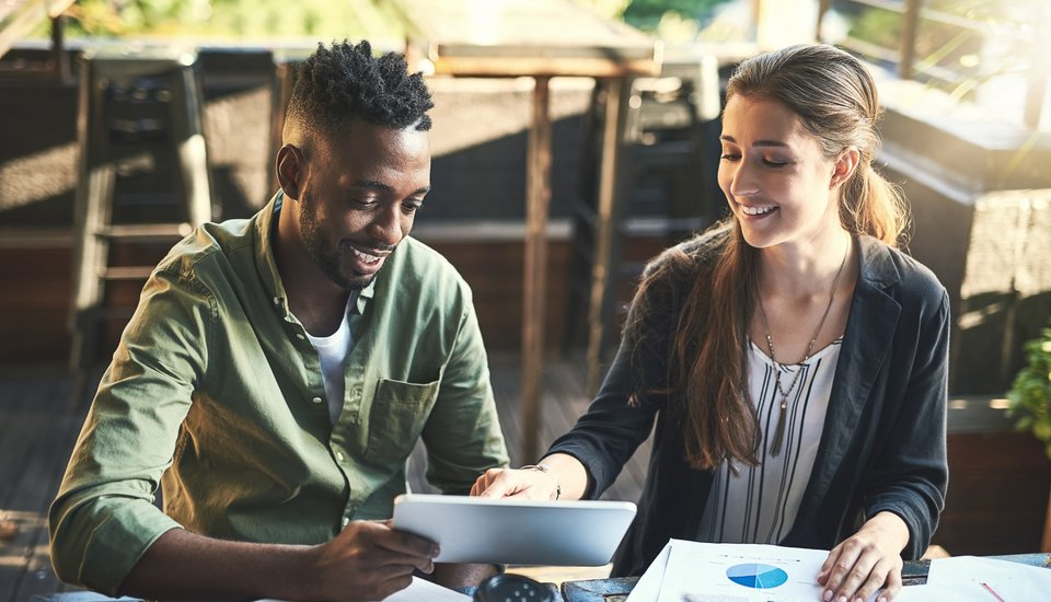 A man and a woman sit at a table with papers and charts, smiling and looking at a tablet together in a bright, casual setting.