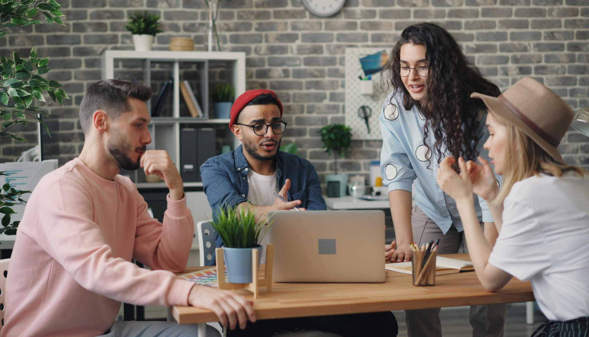 Four young adults sit and stand around a wooden table, engaged in discussion while looking at a laptop. The modern office setting—with shelves, plants, and a brick wall—reflects their sense of community as they connect over ideas.