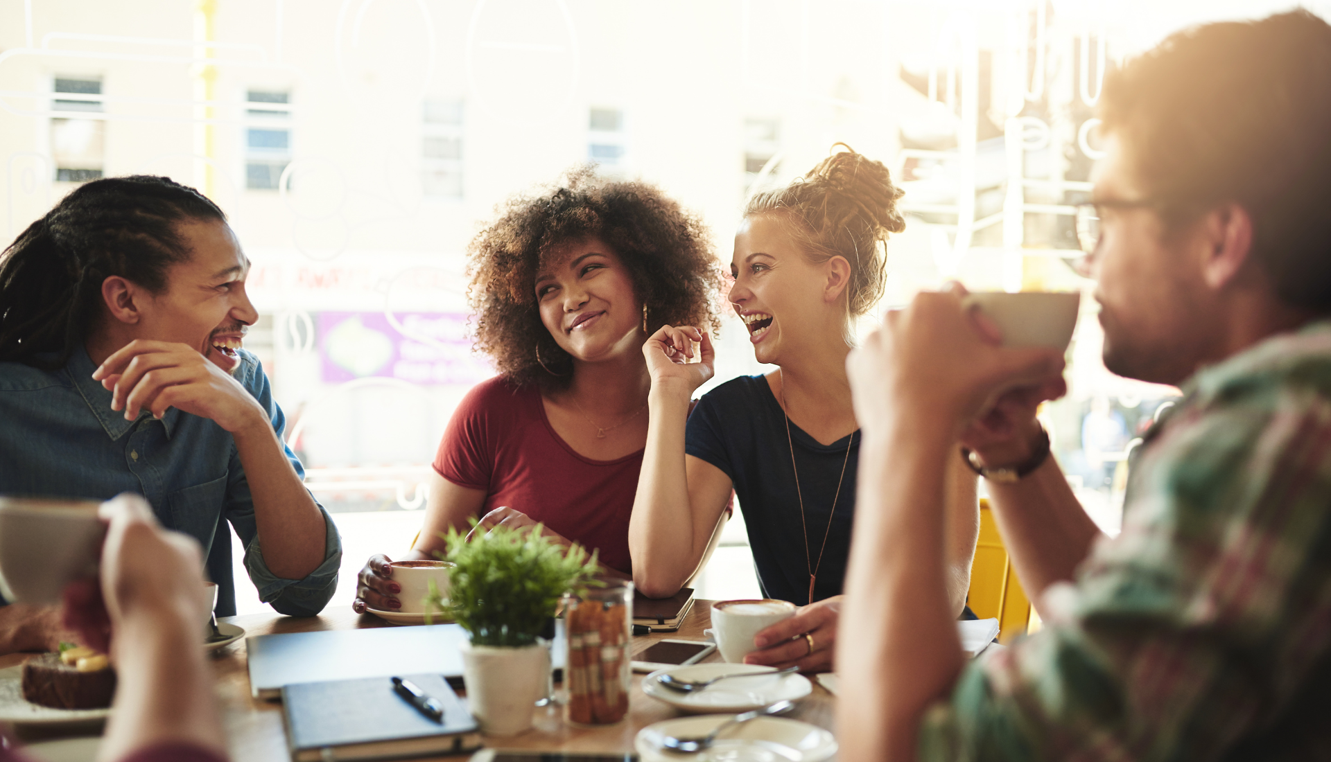 Four people sit around a café table, smiling and laughing together while discussing women's health and AI advancements. Sunlight streams through the window, creating a warm and friendly atmosphere.