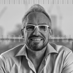 A smiling man with light-colored hair, a beard, and white-framed glasses, wearing an open-collared shirt, sits indoors with a blurred cityscape behind him—capturing the thoughtful mood often found during productive disagreement.