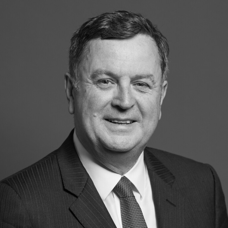 A middle-aged man wearing a dark suit, patterned tie, and light shirt, smiling at the camera against a plain background; the photo is in black and white.