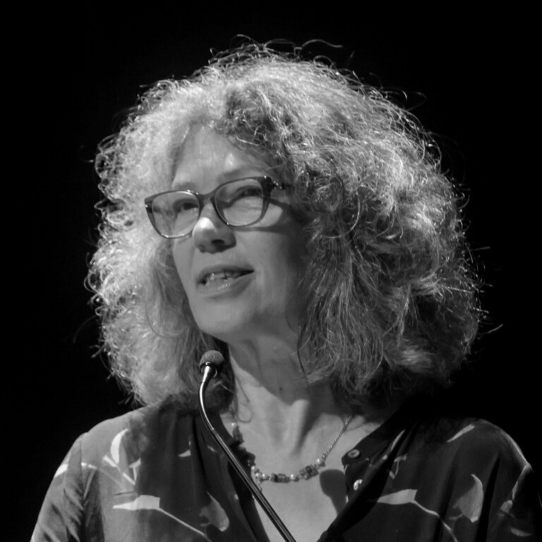 A woman with curly hair and glasses speaks at a microphone, engaging in productive disagreement. She wears a patterned blouse and a necklace, set against a dark background in this black-and-white image.