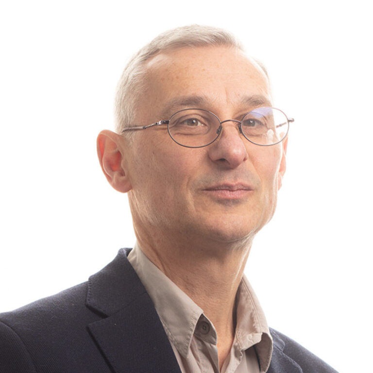 A middle-aged person with short gray hair and glasses, wearing a light-colored shirt and dark blazer, poses against a plain white background.