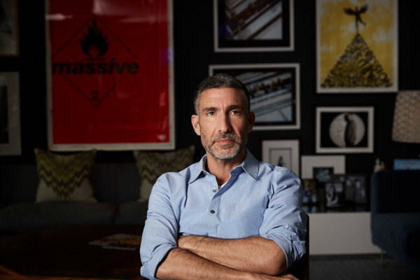 A man with short dark hair and a beard, wearing a light blue shirt, sits with his arms crossed in a room decorated with framed artwork and photographs on the wall behind him.