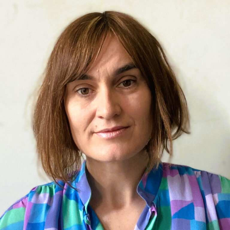 A person with straight, shoulder-length brown hair and light skin is wearing a multicolored, geometric-patterned shirt. They are looking directly at the camera with a neutral expression against a plain background.