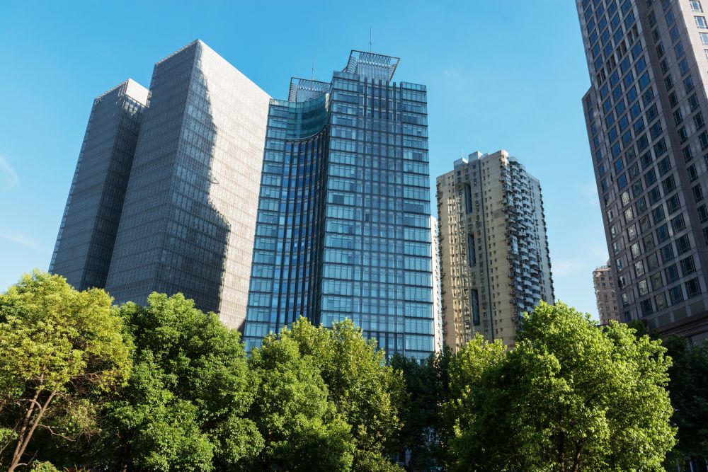 Several modern high-rise office buildings with glass and concrete exteriors stand behind a row of lush green trees under a clear blue sky.