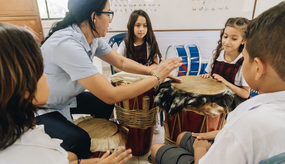 A teacher and young students sit in a circle playing hand drums together in a classroom, focusing on the instruments. The children appear engaged and attentive. A whiteboard and other drums are visible in the background.