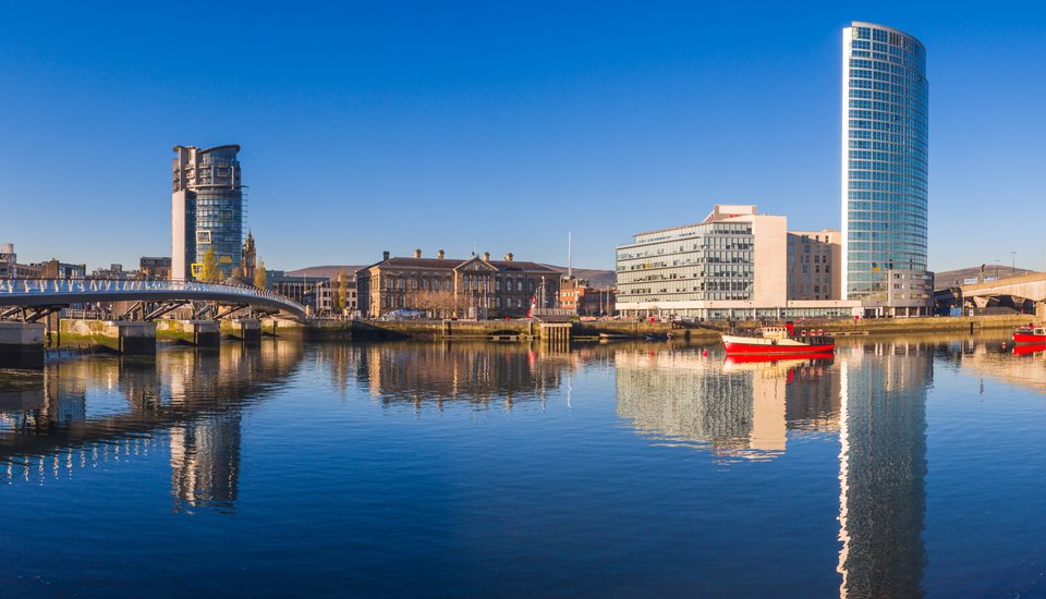 A bright, clear day in Belfast with modern and historic buildings reflected in the calm river, featuring a tall glass skyscraper, red boats, and a pedestrian bridge under a blue sky.