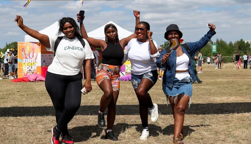 Four women at an outdoor festival pose joyfully, smiling and holding flags to celebrate UK Black Pride. They wear casual summer clothes, with tents and other people in the background under a partly cloudy sky.