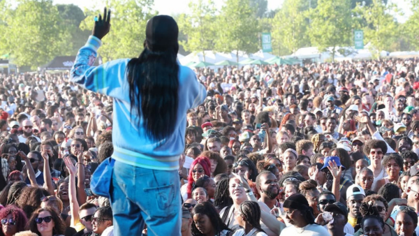 An artist with long hair and a blue jacket performs on an outdoor stage, facing a large, energetic crowd of festival-goers under sunny skies, with green trees and tents in the background.