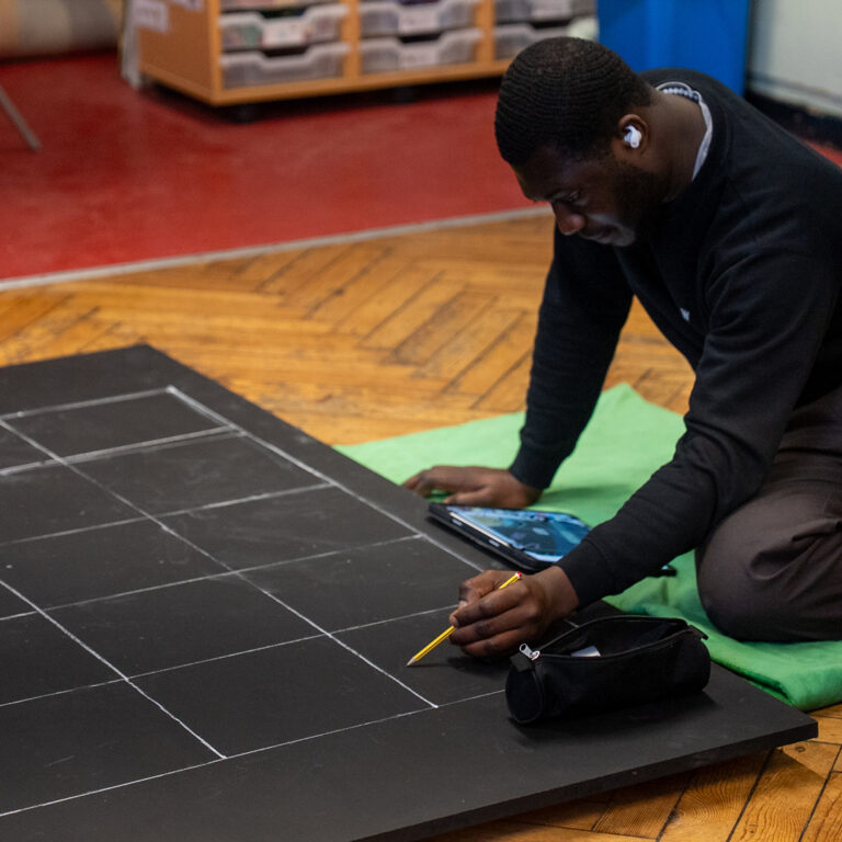 A person sits on a green mat, using a pencil and ruler to draw lines on a large black grid board. They hold a tablet and wear an earbud, with a pencil case nearby on the board.