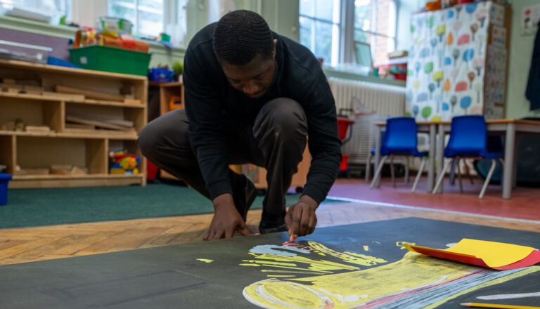 A person kneels on a classroom floor, drawing a colorful picture on a large sheet of black paper. Brightly colored chairs and shelves with toys are visible in the background.