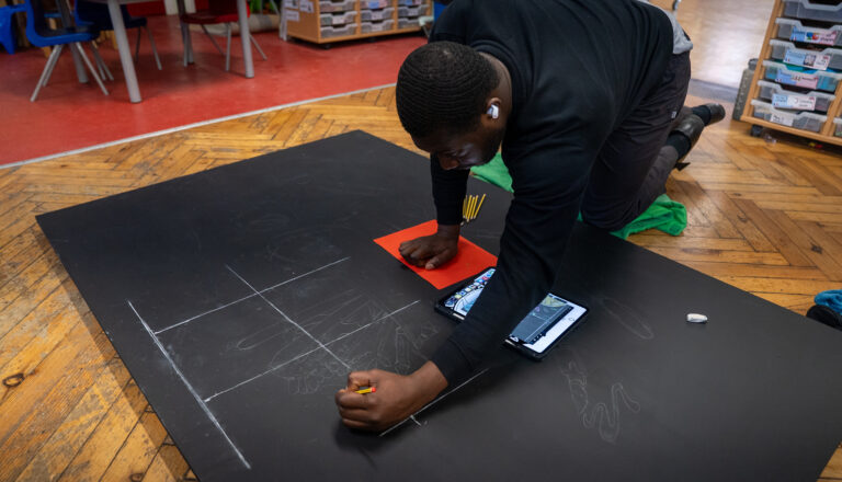 A person kneels on a wooden floor, sketching lines and shapes with a pencil on a large black sheet. An eraser, phone, red notebook, and pencils are beside them. Shelves and colorful chairs are in the background.