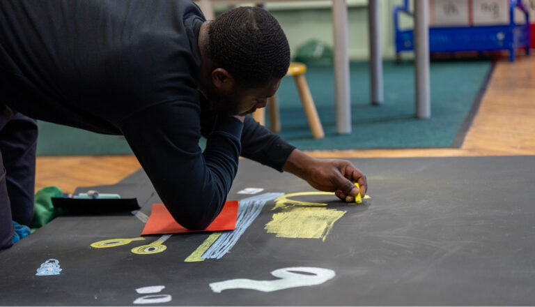 A person is kneeling on the floor, drawing colorful shapes and numbers with chalk on a large black sheet of paper. There are tables, chairs, and a carpeted area in the background.