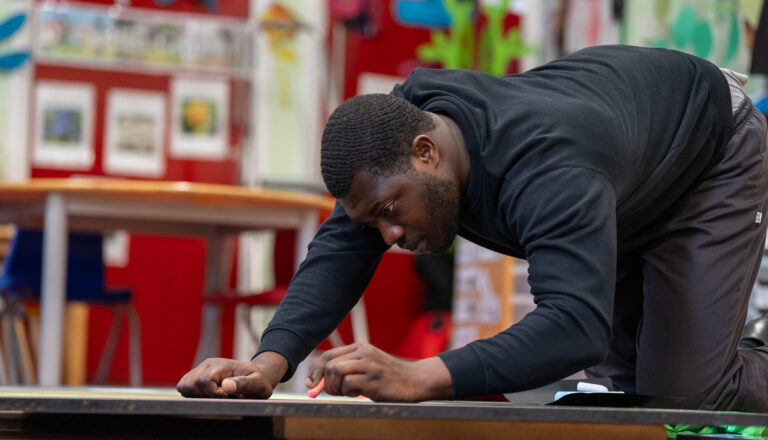 A man in a black hoodie kneels on the floor, focused on measuring or marking a large black surface with a pencil in a colorful classroom or workshop setting.