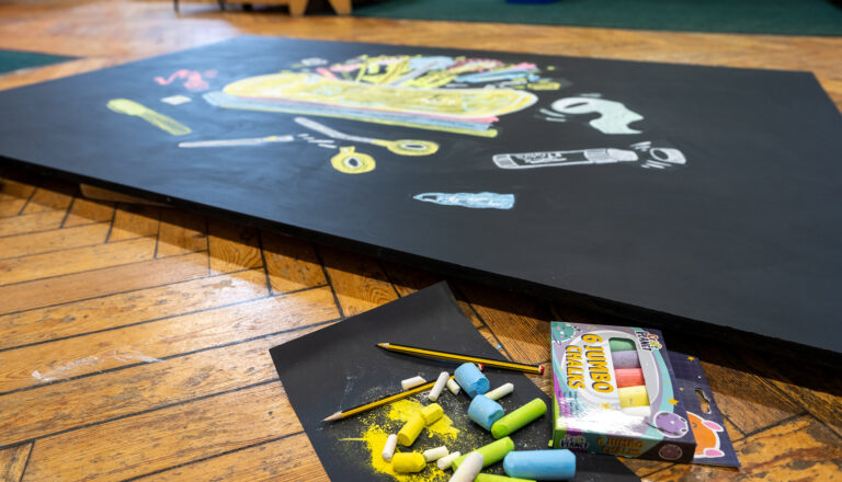 A blackboard with colorful chalk drawings of art supplies lies on a wooden floor. Nearby are scattered jumbo chalk sticks, pencils, and an open box labeled “Jumbo Chalks.”.