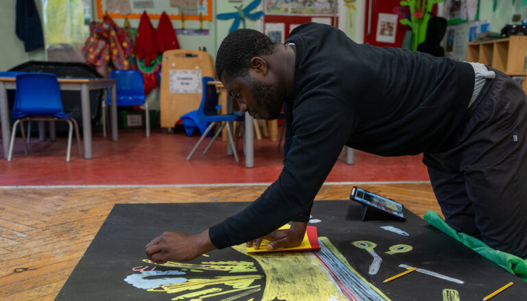 A man kneels on the floor, drawing with chalk on a large black paper. He is surrounded by art supplies and classroom furniture, working intently on a colorful, creative project in a classroom setting.
