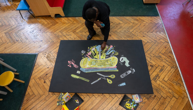A person draws colorful, imaginative objects and shapes with chalk on a large black paper placed on a wooden floor in a classroom setting; art supplies are scattered nearby.