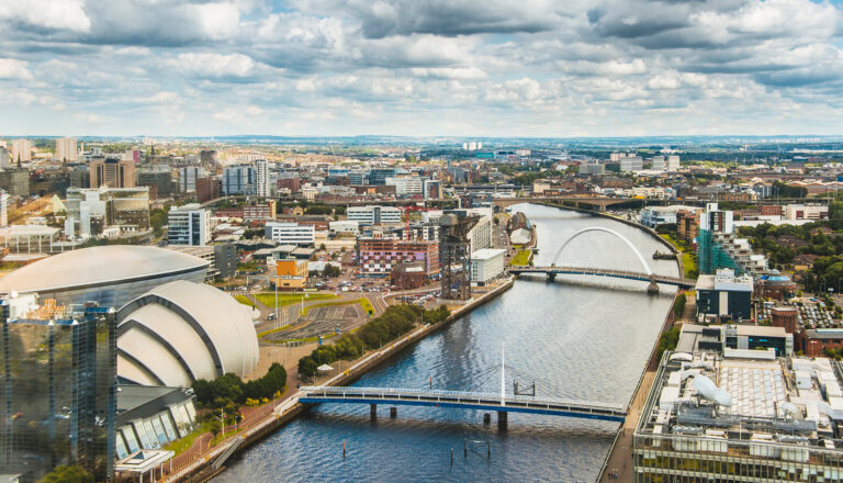 Aerial view of a city with modern buildings, bridges, and a river flowing through the center under a cloudy sky. The scene includes distinctive domed and curved structures along the waterfront.
