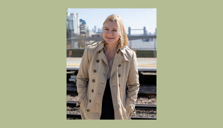 A person wearing a beige trench coat stands outdoors by train tracks, smiling. A city skyline and bridge are visible in the background under a clear sky.