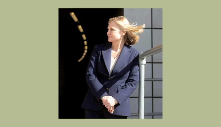 A woman in a navy blazer stands by a railing, looking to the side with sunlight illuminating her face and hair, set against a dark tunnel entrance and tiled wall background.