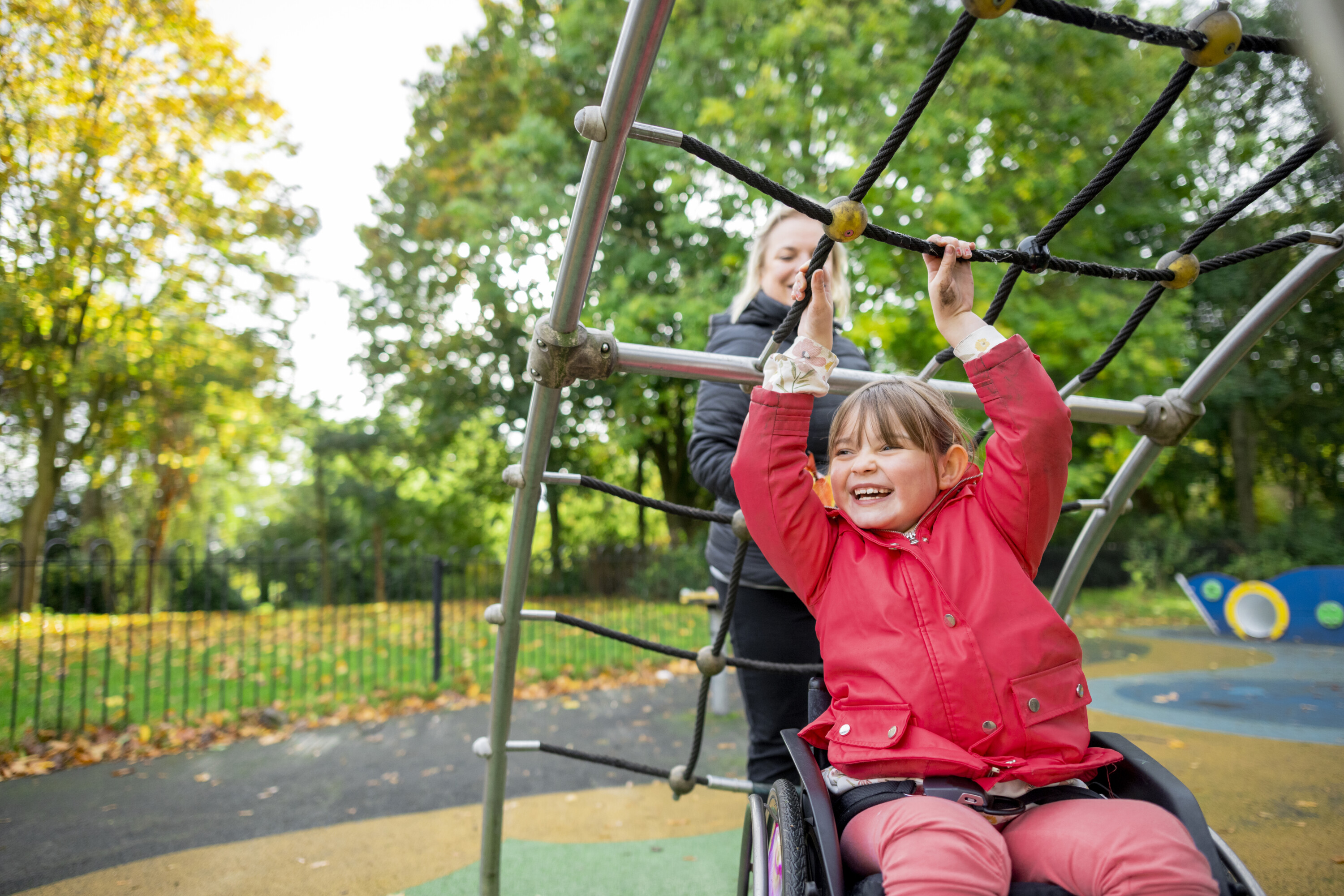 A young girl in a wheelchair smiles as she hangs from playground climbing bars, with an adult behind her. The playground is outdoors, surrounded by green trees and autumn leaves.