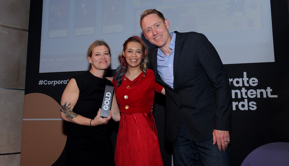 Three people smile at the camera; the woman on the left holds a gold award trophy. They stand before a backdrop reading "Corporate Content Awards," celebrating excellence in corporate storytelling and digital content.